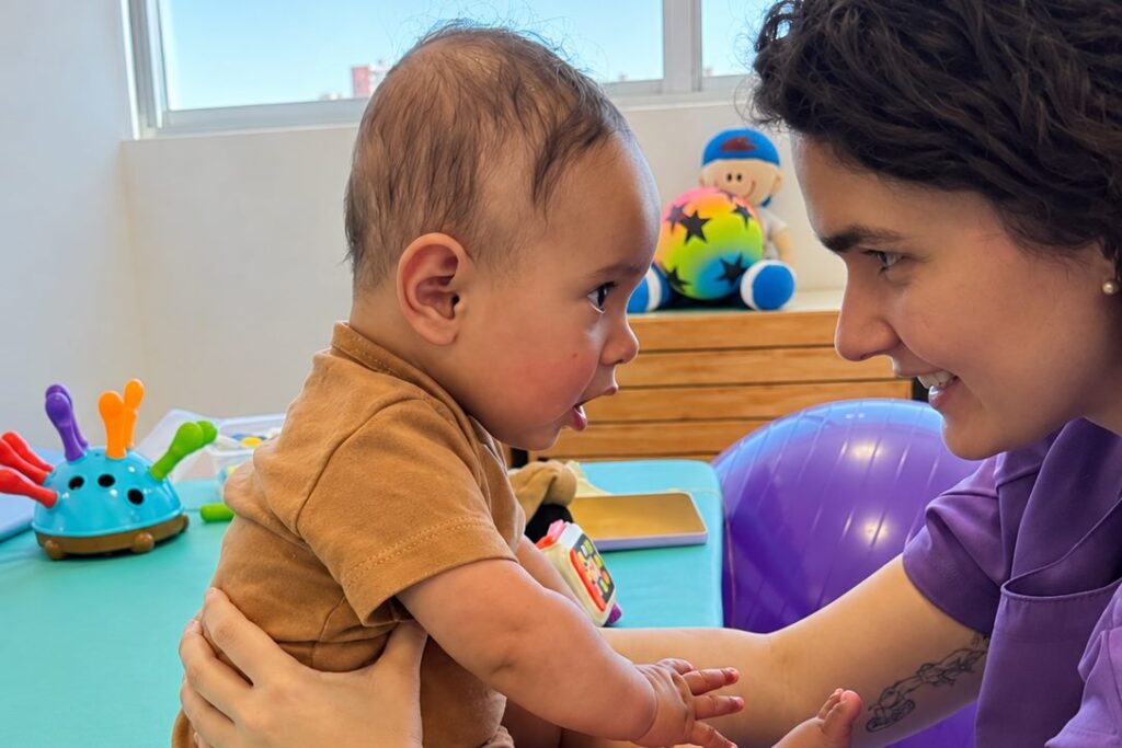 Bebê sentado com apoio na maca, de frente para uma fisioterapeuta sorridente, durante avaliação de fisioterapia infantil em Porto Alegre
