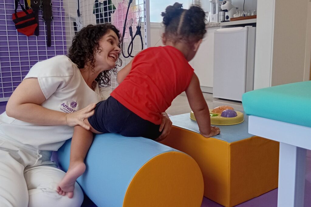 Fisioterapeuta da AmarElo sorri ao apoiar uma criança que sobe em um rolo e em um bloco de espuma durante sessão de fisioterapia neuropediátrica em Porto Alegre